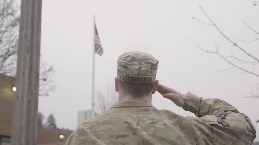 A military man dressed in army fatigues saluting outside towards the american flag