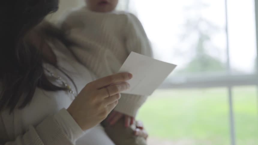 A pregnant mom holds her son and shows him a photograph while standing near a window