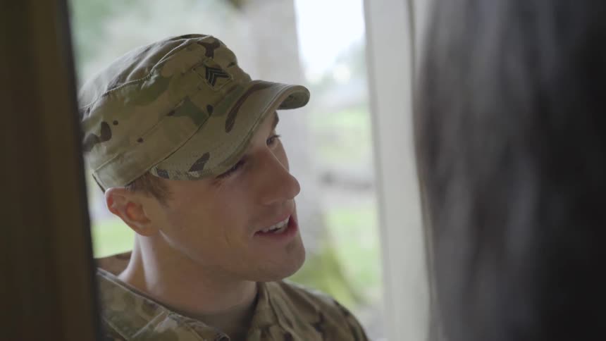 A soldier in a camo battle dress uniform smiles and talks to a woman with a baby while standing in a doorway