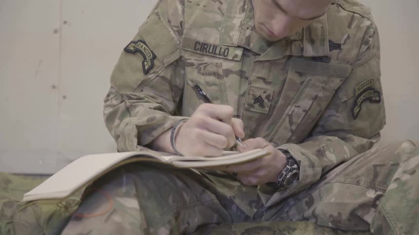 A man in a military outfit writing in a notebook while sitting on the floor