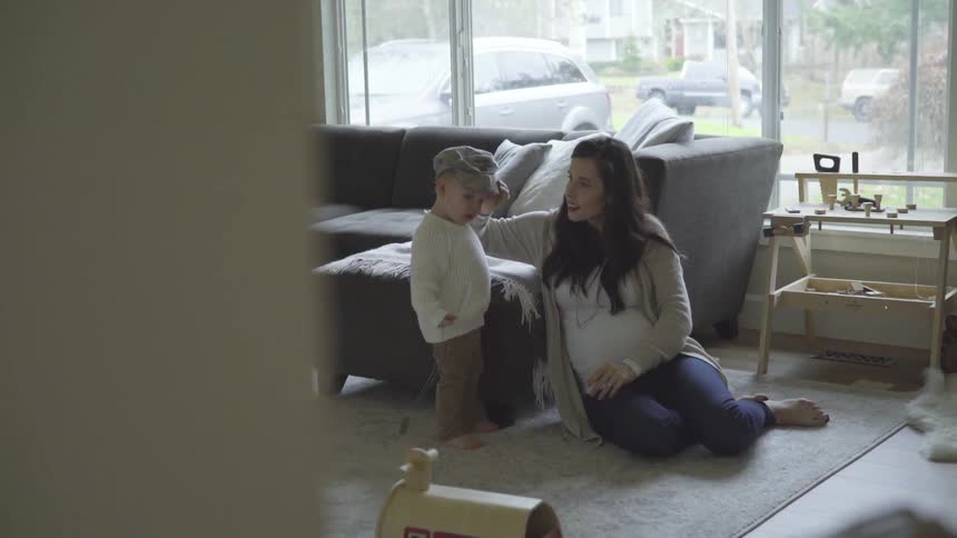 A pregnant mom sitting on the rug in the living room puts a cap on her young sons head