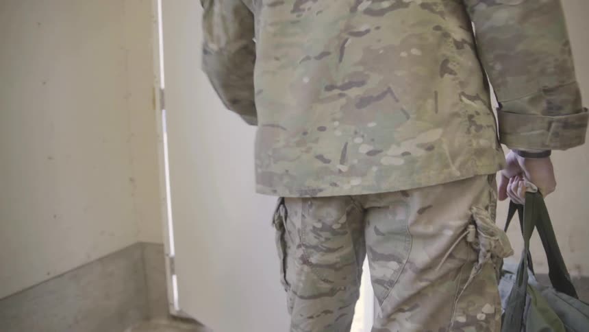 A man wearing military gear leaves a building holding a bag
