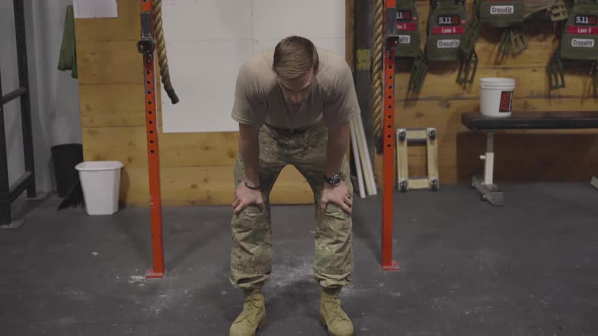 A man wearing a battle dress uniform and boots bends over with hands on knees to catch his breath after a workout