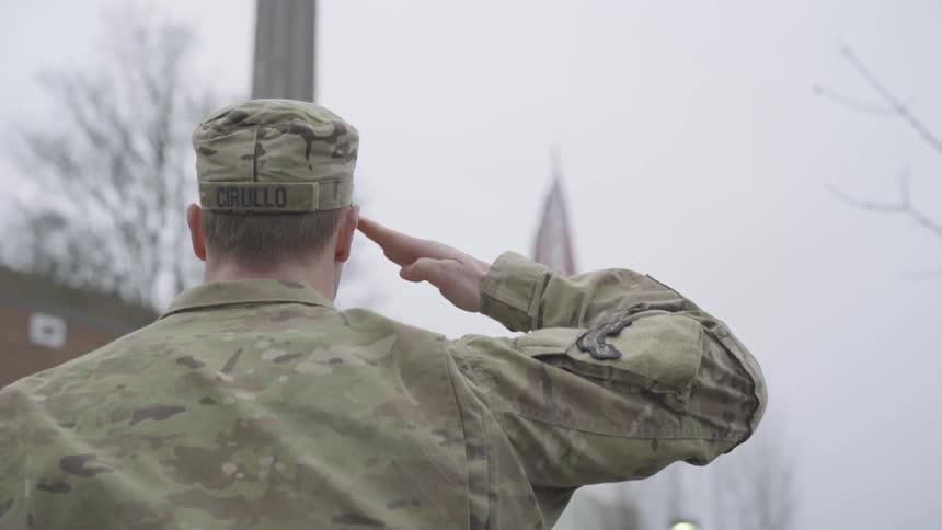 On an overcast day a large man in a camouflage hat and jacket stands and salutes and american flag waving in the distance