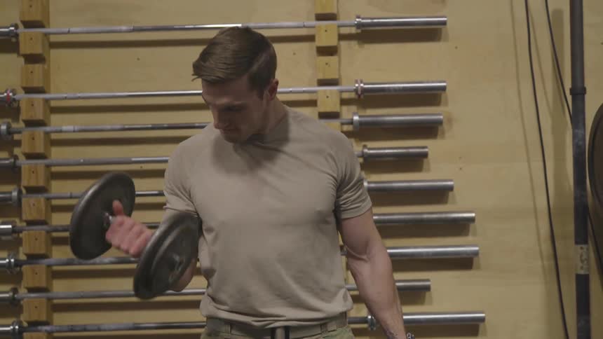 A man wearing a t shirt and a battle dress uniform does bicep curls with hand weights