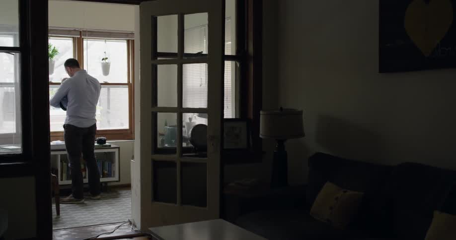 A man stands in a home holding a baby and looking out some windows