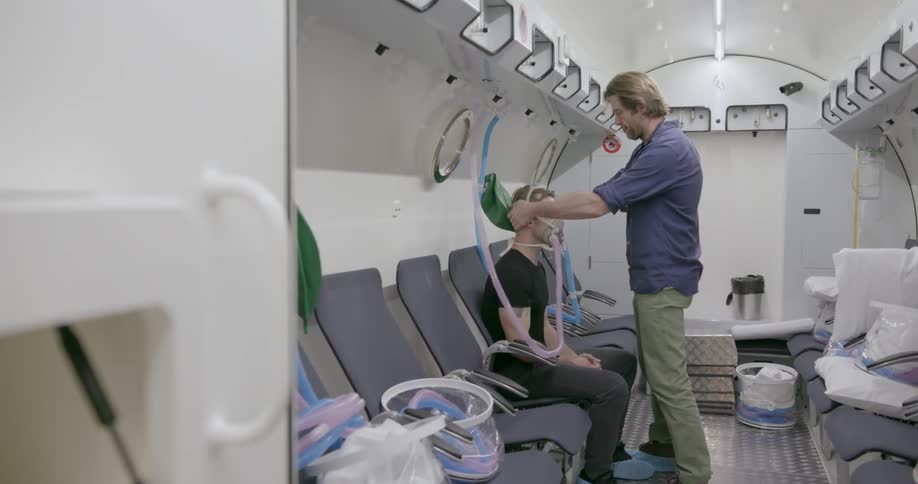 A man puts an oxygen mask on another man that's sitting in front of him