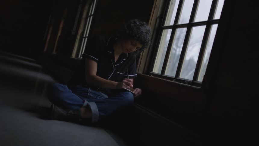 A woman with short curly hair sits on the floor of a long dark hallway and writes in a small notebook next to a large window
