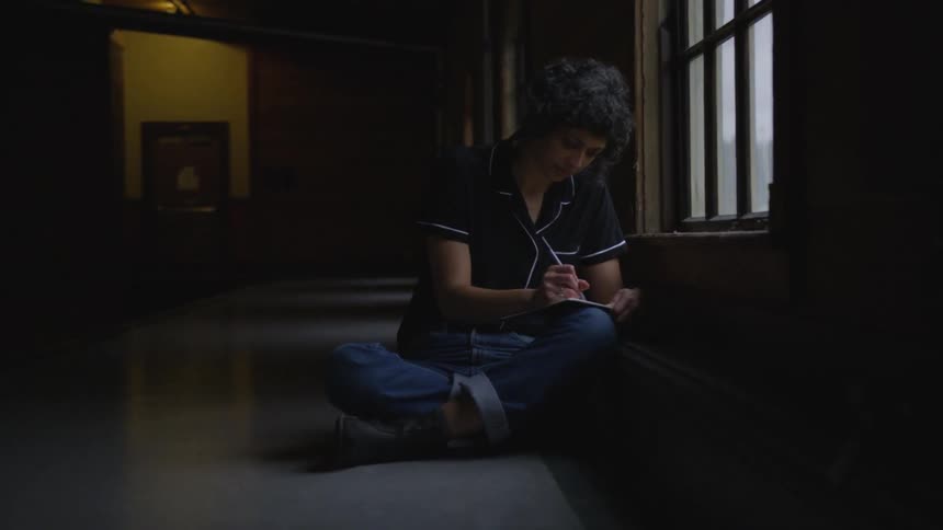 A woman sitting on the floor cross legged near a low window while writing on a notepad