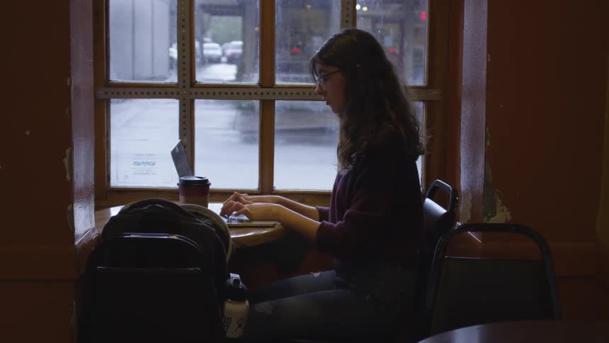 Woman sits in front of a window typing on a laptop