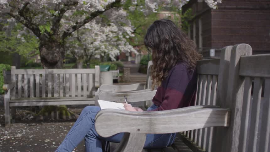 A woman sits on a wooden bench in a garden writing in a notebook