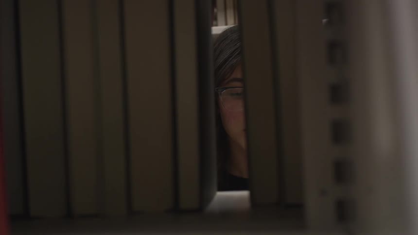 A woman becomes visible when she removes a book from a shelf in a library