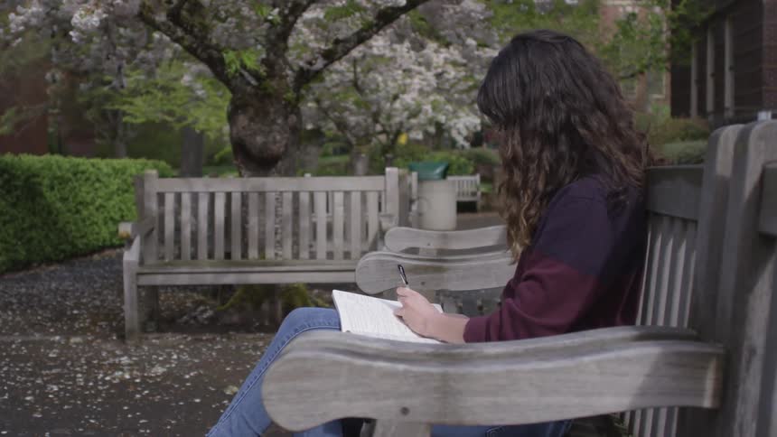 A young brunette woman writing in a notebook while sitting on a wooden bench outside during the day