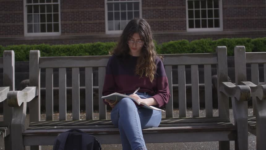 A woman is sitting on a bench outside writing in a notebook