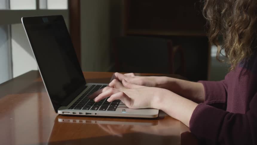 A woman in a purple sweater types on her laptop at a wooden desk