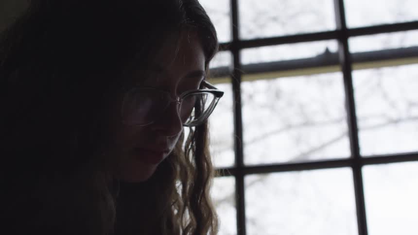 A girl smiles as she looks down and reads near a large window