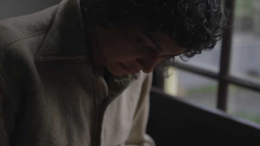 A young woman with dark curly hair sits and concentrates on something while she wears a thick warm coat next to a large window