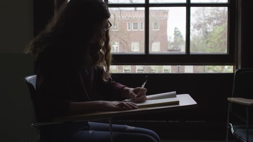 A female student sitting at a desk and writing in a notebook in a dark classroom during the day