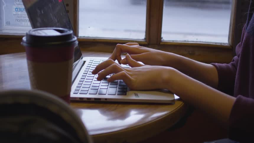 A woman sits in a cozy coffee shop at a small table next to a window as she types on her laptop computer