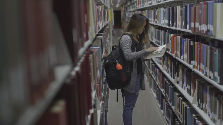 Woman pages through a book while standing in a library corridor