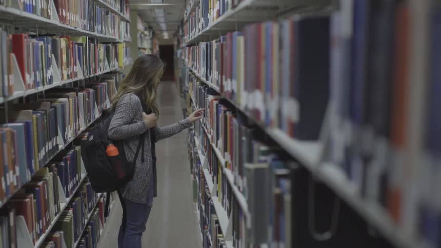 A young woman with a backpack and a gray coat stands amidst long rows of books in a library