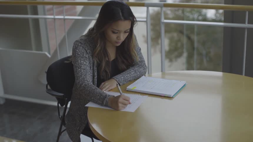 A woman sits at a table near large windows and writes on a piece of paper from her notebook