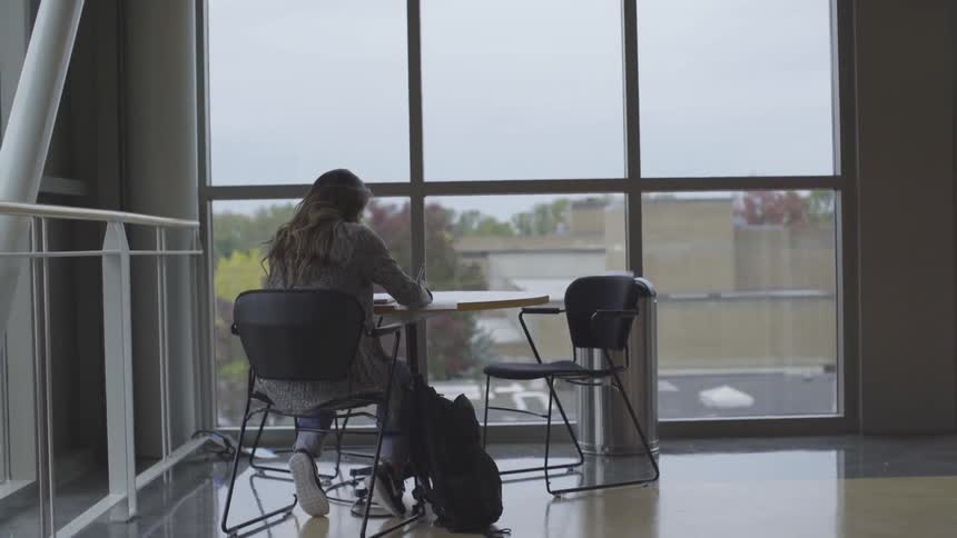 A woman sitting at a table in front of a large window writing on a sheet of paper