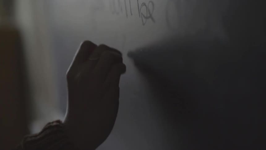 A person wearing a ring writing on a chalkboard in a dim room during the day