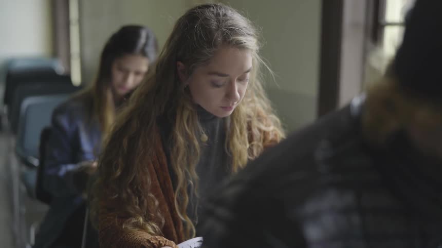 A young woman seated near her peers in a classroom looks down at book that she has open on her desk