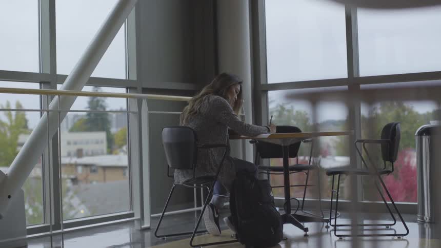 A woman sits writing at a table in the corner with large windows on either side