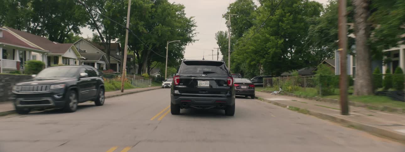 A dark colored vehicle traveling through a neighborhood on a cloudy day