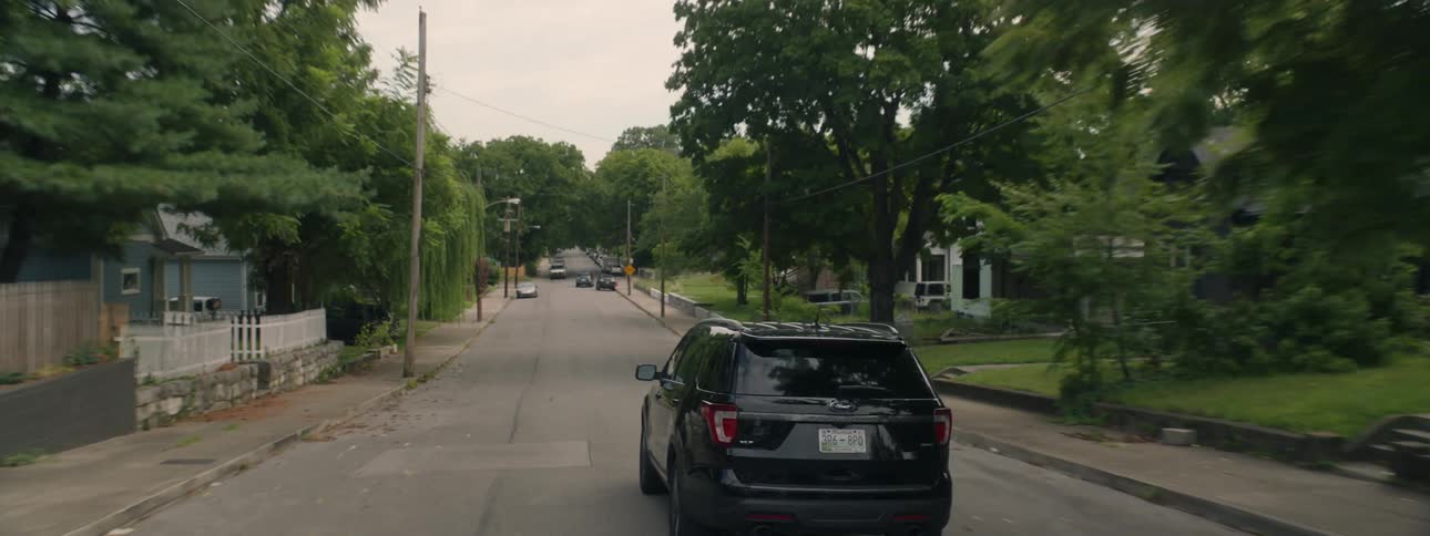 A darkly colored vehicle traveling down a road in a residential area during the day