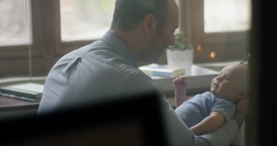 A man holds a baby at a desk by a window