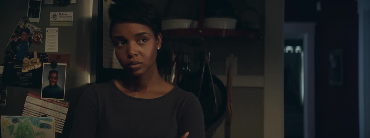 A woman leaning against a refrigerator with arms crossed in front of her looks up then back down again
