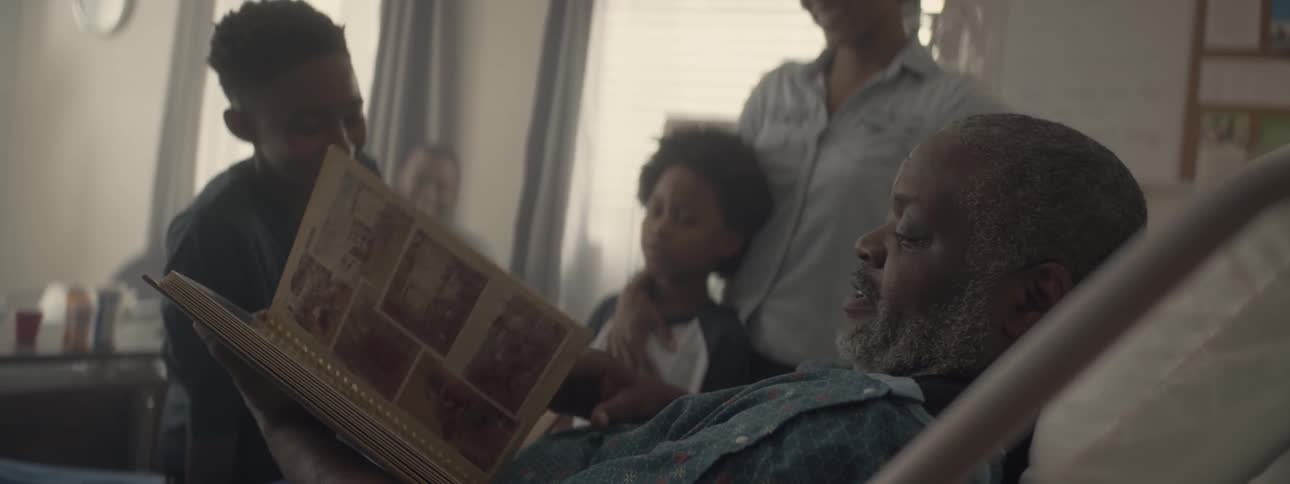 Two adults and two children are visiting with a man in a hospital bed who is looking through an old photo album