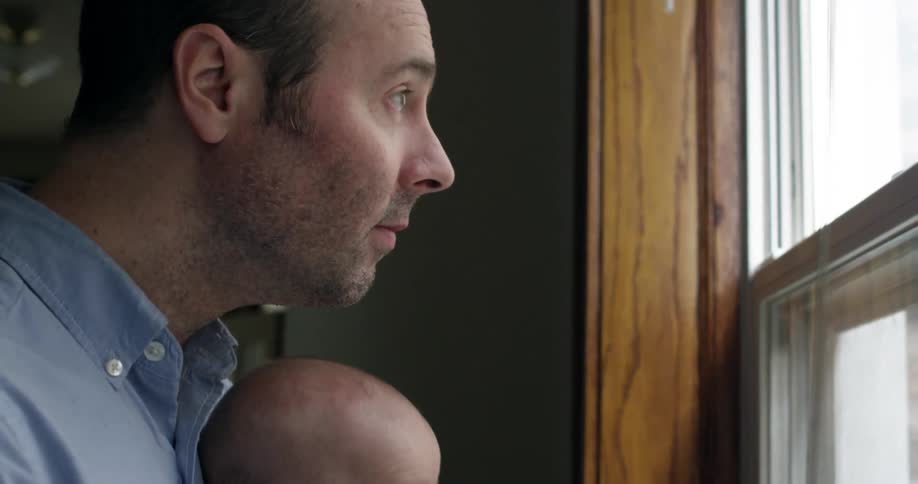 A man holds a baby and looks out a window