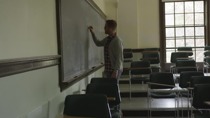 Man in glasses writes on chalkboard with chalk in classroom by day
