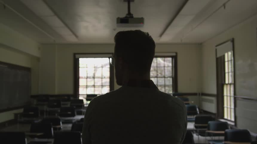 A man wearing glasses stands in an empty classroom as he looks around