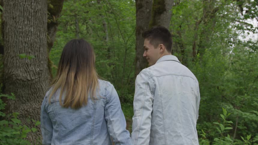 A teenage boy and a teenage girl walk through a forest holding hands
