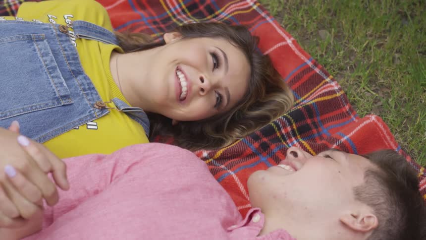 Young woman and young man lay on a blanket holding hands and smiling