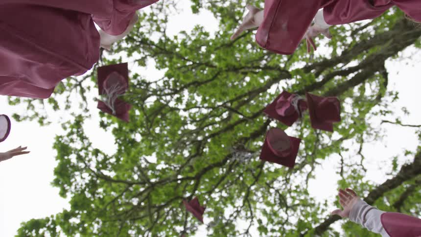 Graduates throw their caps into the air underneath a tree