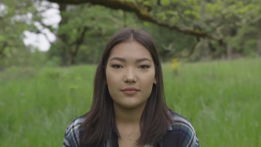 A girl with straight brown hair sits in a field of tall grass with trees and branches behind her