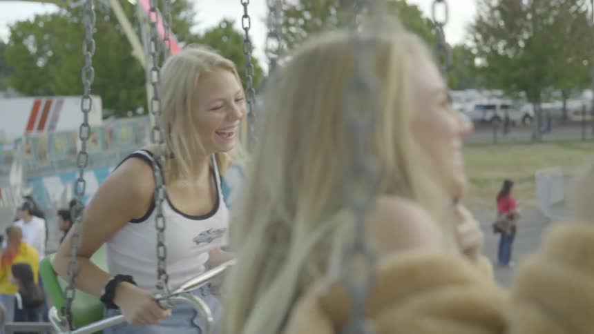 Two blond teenage friends laugh as they sit on carnival ride swings above the fairgrounds