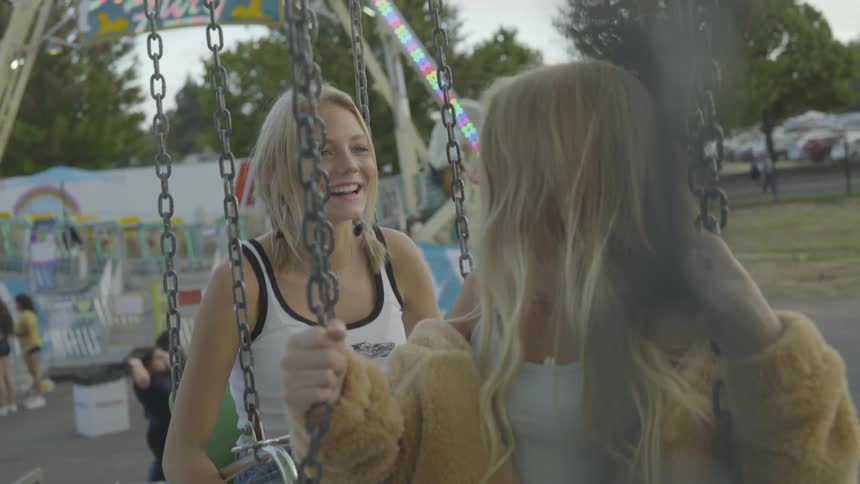 Two girls sit on a swing at a fair smiling and talking