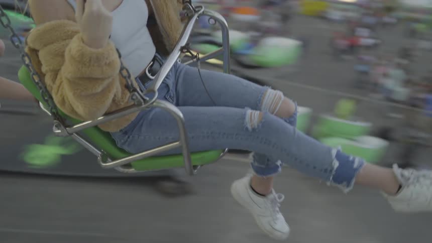 A female wearing a fur jacket and holey jeans is swinging her legs as she is flying in a swing at an amusement park