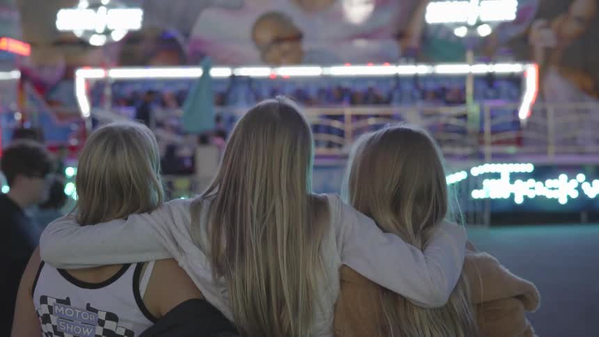 Three blonde women stand with their arms around one another facing a fairground ride