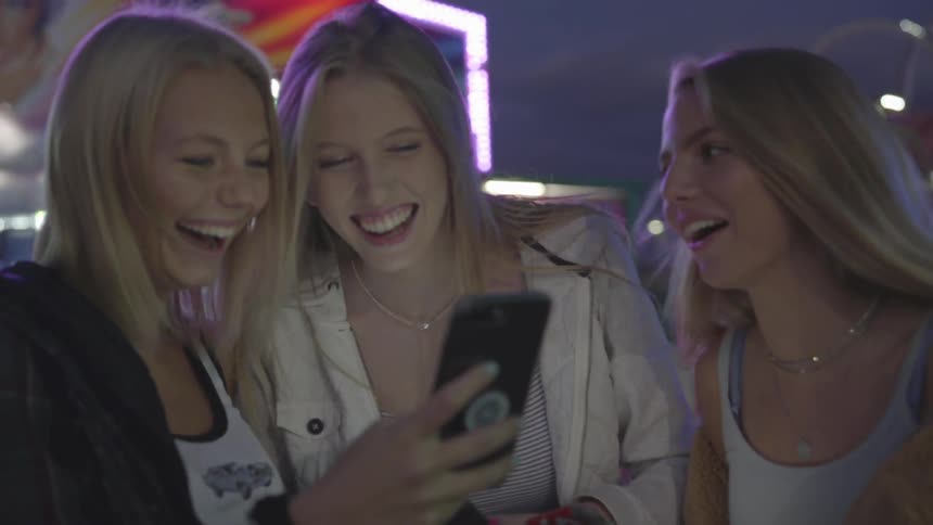 Three blonde women look at a phone and laugh