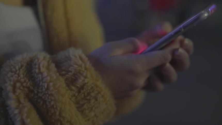 A woman in a fuzzy coat scrolls on a smartphone while standing outside in a crowd of people
