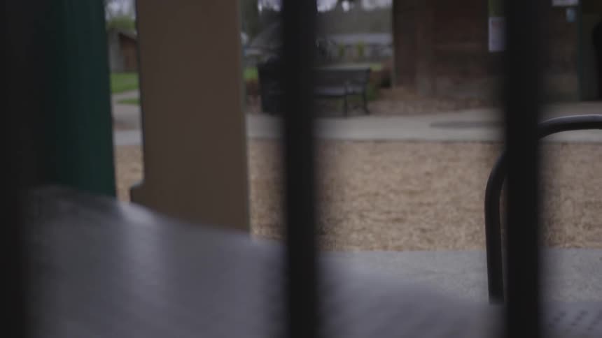 An empty park and bench is viewed through the bars of a playground structure