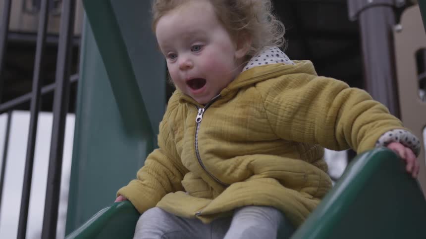 Toddler who has blue eyes and wearing yellow coat goes down the green slide on a cloudy day